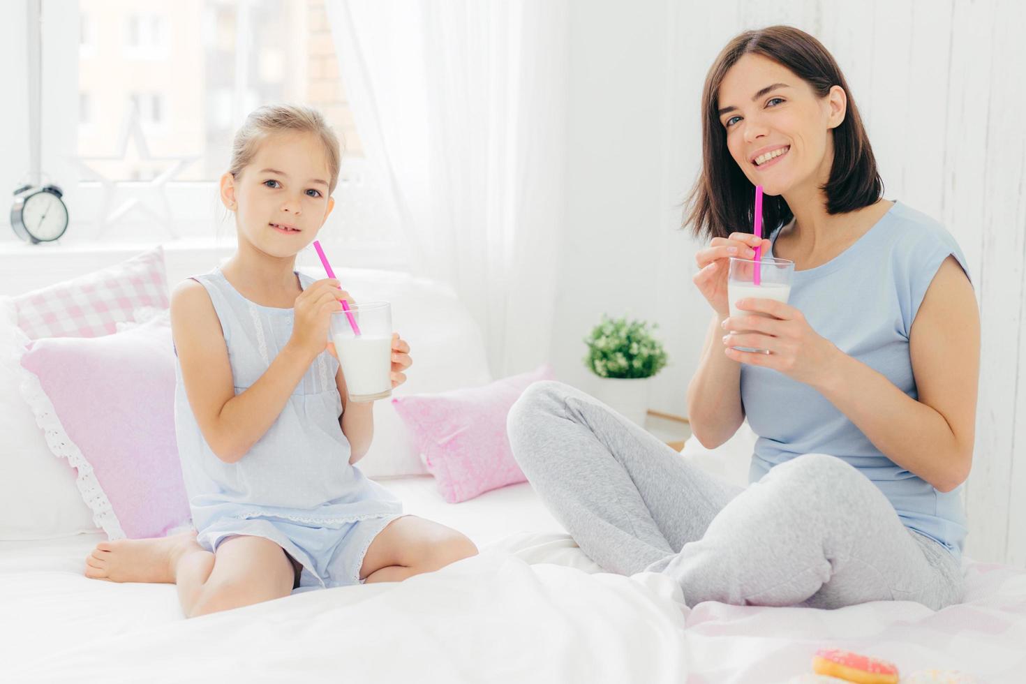 la giovane mamma bruna felice e la sua bella figlia si siedono sul letto, bevono frullati e mangiano deliziose ciambelle, si godono la calma atmosfera domestica in camera da letto. persone, tempo libero, biancheria da letto, concetto di maternità foto