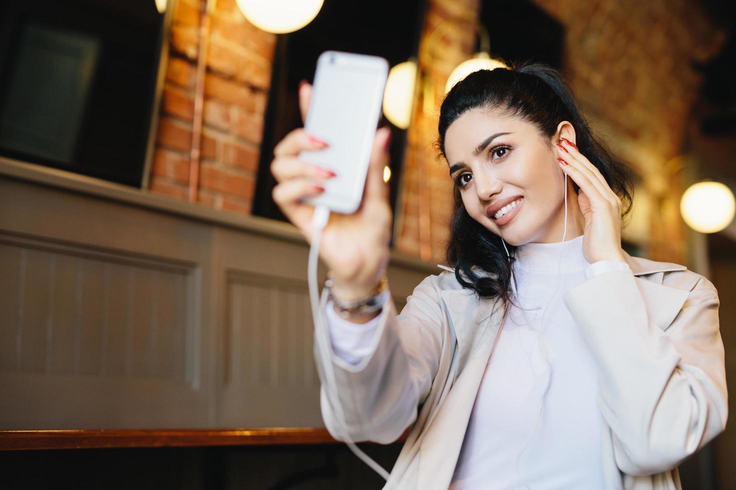 donna attraente con capelli scuri, pelle sana e occhi luminosi che tiene lo smartphone in mano facendo selfie e ascoltando musica o audiolibro mentre si riposa nella caffetteria. concetto di persone e tempo libero foto