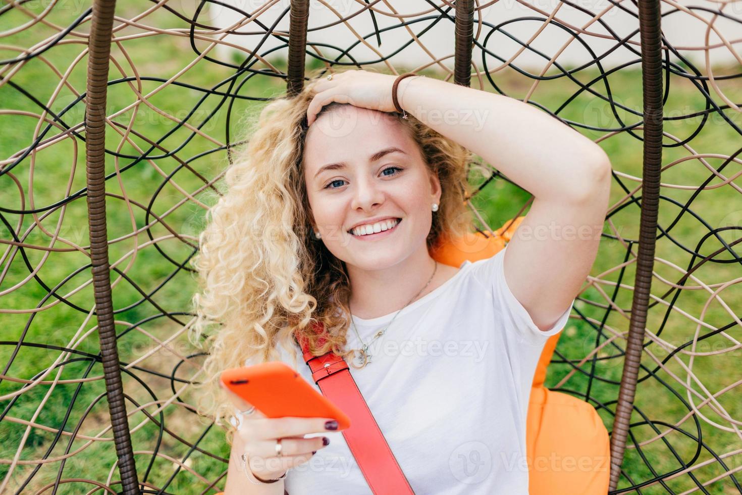 donna europea sorridente con ampio sorriso, capelli ricci, vestita con una maglietta bianca casual, utilizza un moderno dispositivo elettronico per navigare sui social network, felice di ricevere un messaggio di invito alla festa foto