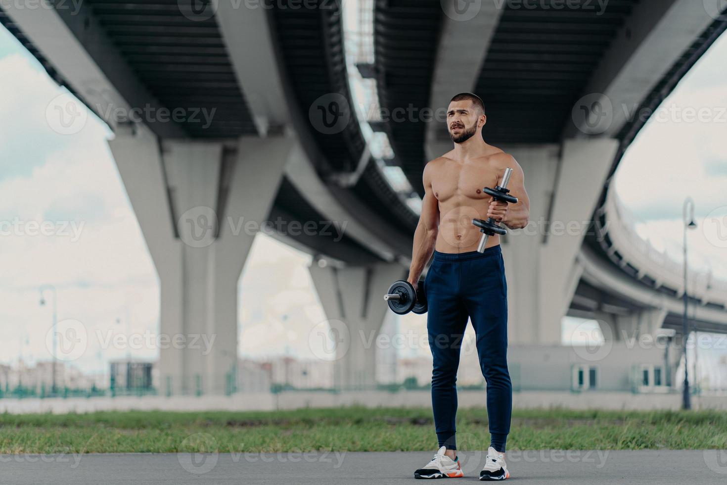 muscoloso atletico bodybuilder barbuto solleva manubri lavora sui bicipiti ha un duro allenamento vicino al ponte vestito con pantaloni sportivi e scarpe da ginnastica, essendo motivato, ama lo sport. concetto di stile di vita sano foto