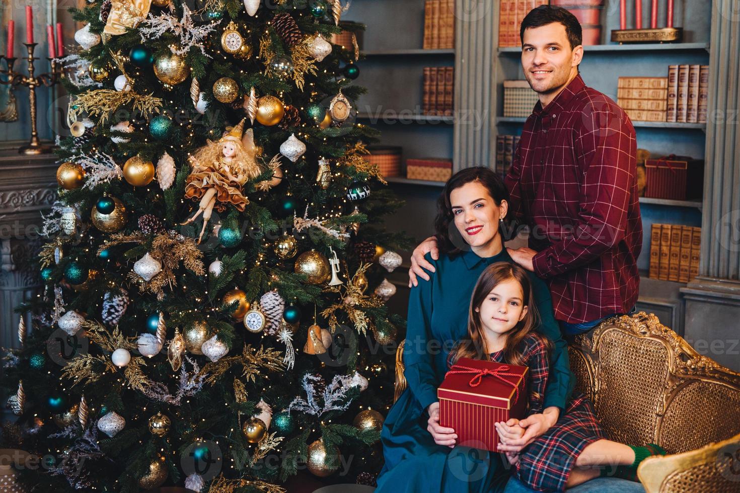 bella donna bruna abbraccia la sua piccola figlia e suo marito si siedono insieme vicino all'albero di Capodanno decorato, essendo felice di trascorrere le vacanze nella cerchia familiare. famiglia affettuosa con sguardi felici foto