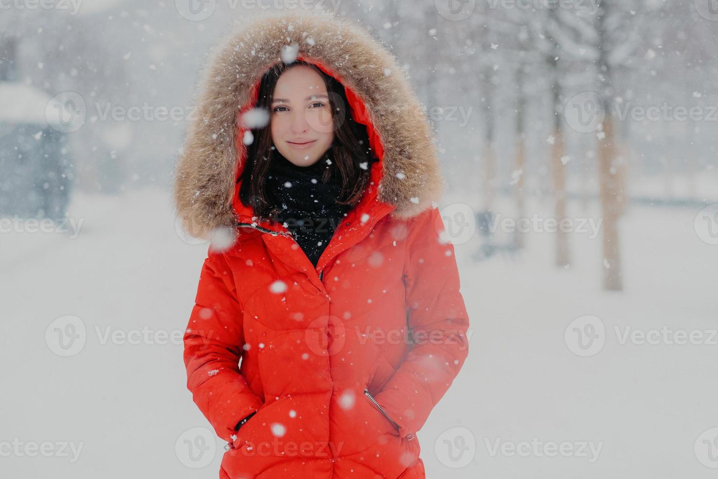 inquadratura alla vita di una donna sorridente attraente con pelle sana, capelli scuri, indossa una giacca rossa, tiene le mani in tasca durante il gelo, si trova su uno sfondo bianco di neve, gode di forti nevicate foto