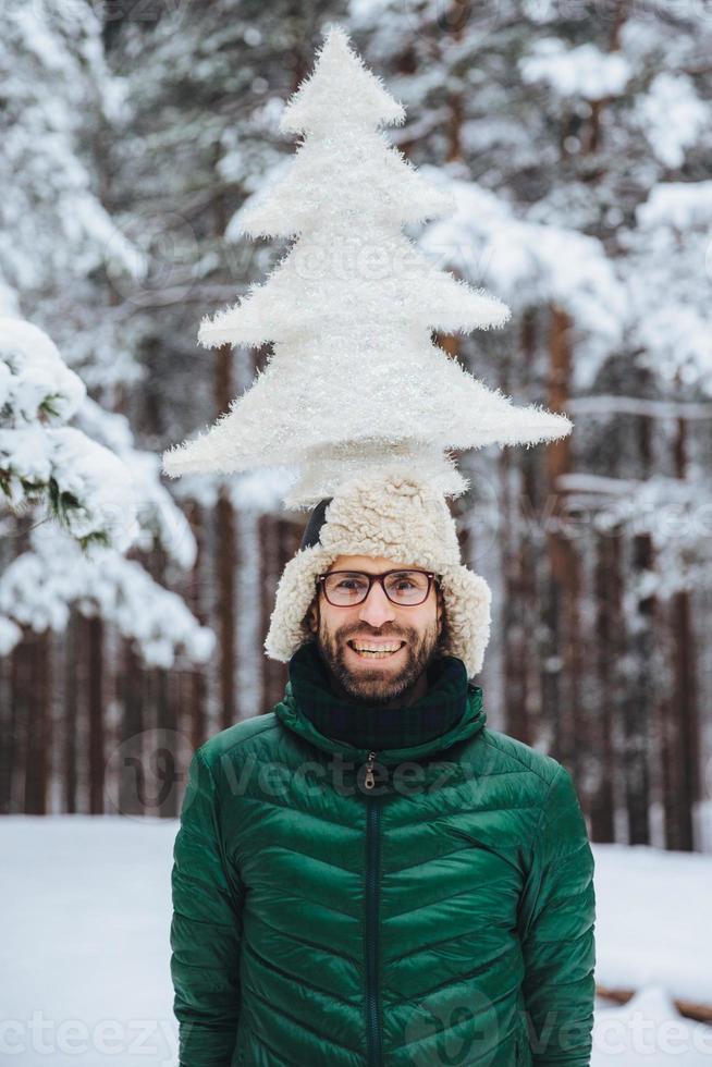 ritratto verticale di allegro uomo barbuto si diverte da solo nella foresta invernale, mantiene un abete artificiale, posa all'aperto, ammira il gelido clima nevoso, esprime positività ed emozioni piacevoli foto