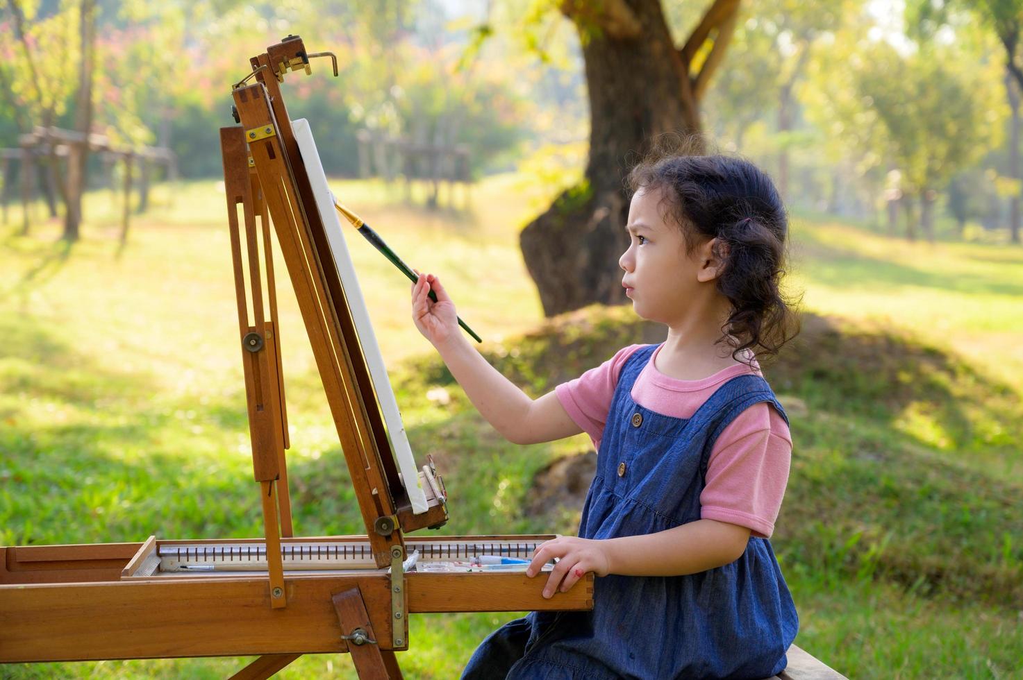 una bambina è seduta sulla panca di legno e dipinta sulla tela posta su un leggio da disegno foto