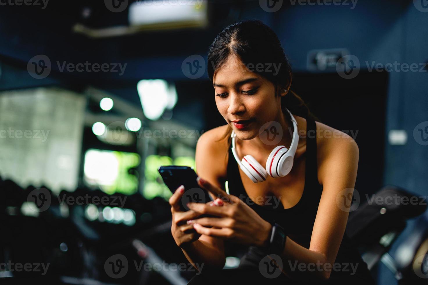 ragazza in palestra che si esercita usa un telefono cellulare ascoltando musica con cuffie over-ear bianche e utilizzando un timer digitale del battito cardiaco. sono strutturati esercizi di esercizio sistematici. foto