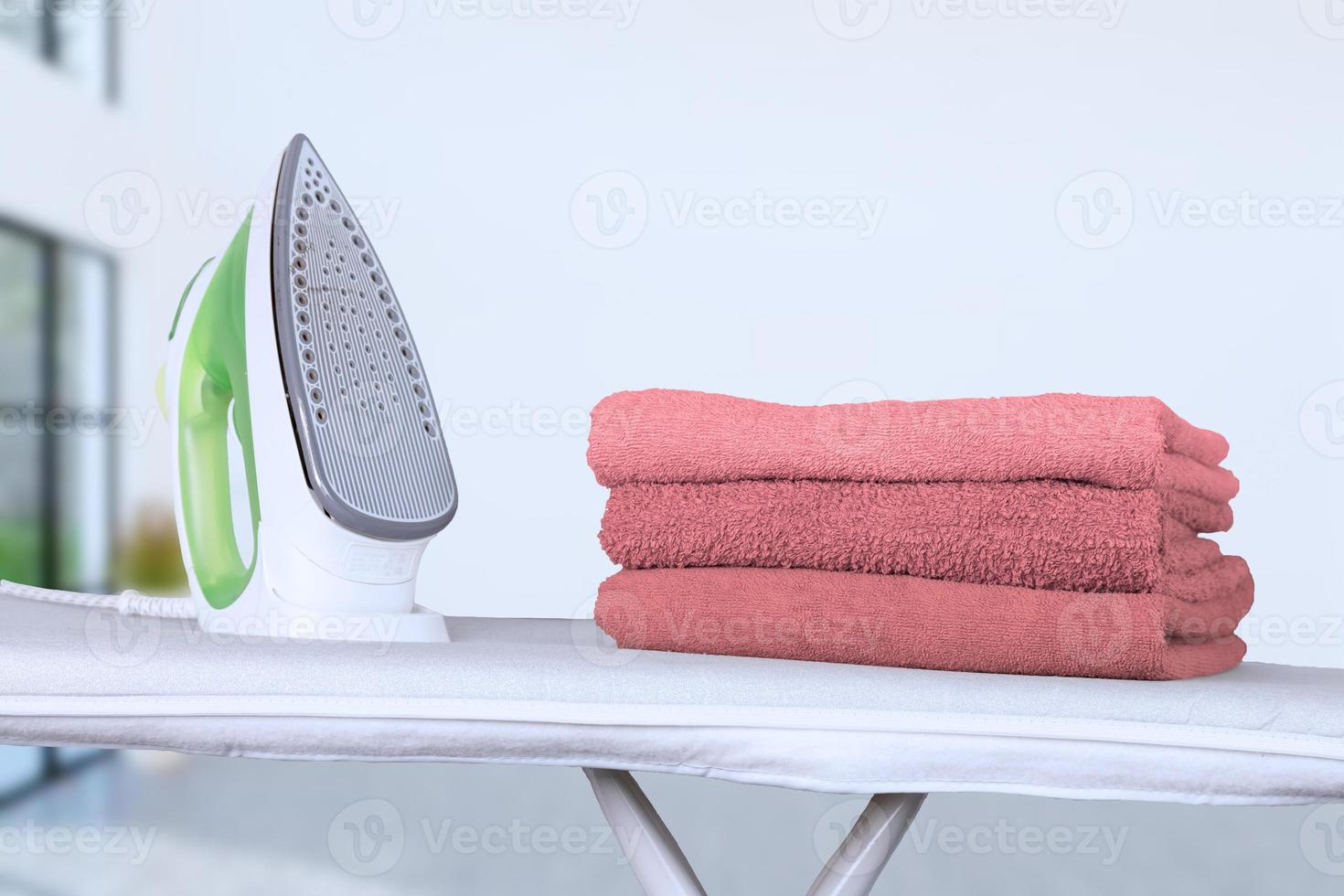 un ferro da stiro elettrico poggia su un'asse da stiro e asciugamani da bagno rosa piegati sullo sfondo di un interno di casa. sfondo sfocato, messa a fuoco morbida. foto