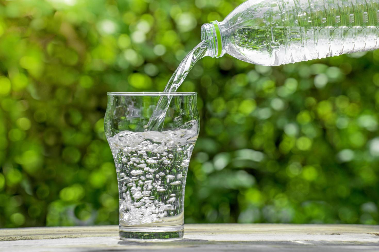 versare l'acqua da una bottiglia di plastica in un bicchiere. lo sfondo delle piante in giardino. focalizzazione morbida. foto