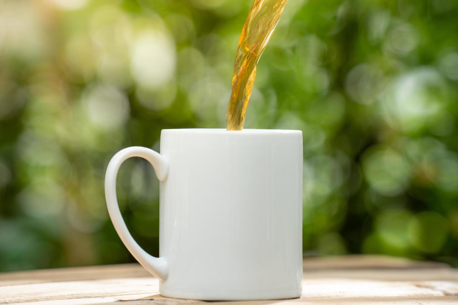 tè versato in una tazza da caffè in ceramica bianca sul pavimento di legno, sfondo bokeh albero verde. effetto messa a fuoco morbida. foto