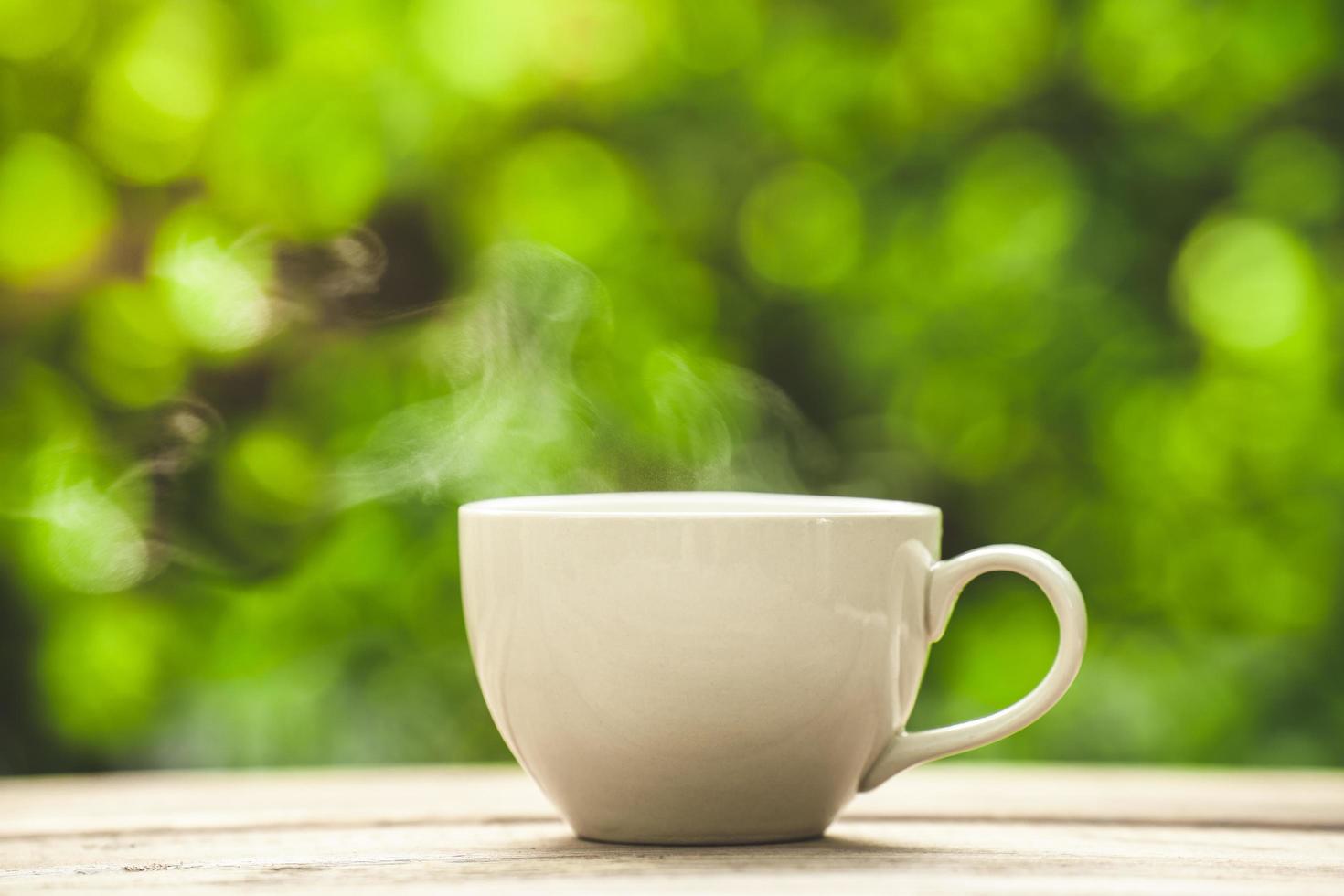 tazza da caffè in ceramica bianca sul pavimento di legno, sfondo bokeh albero verde. effetto messa a fuoco morbida. foto