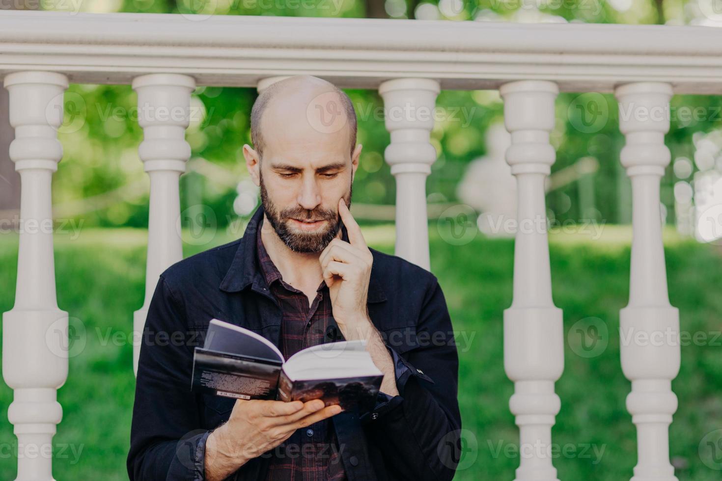 persone, stile di vita, educazione, concetto di hobby. attraente maschio di mezza età con stoppia scura, tiene un libro, si concentra sulla pagina, legge informazioni informative, posa all'aperto, ha un'espressione seria. foto