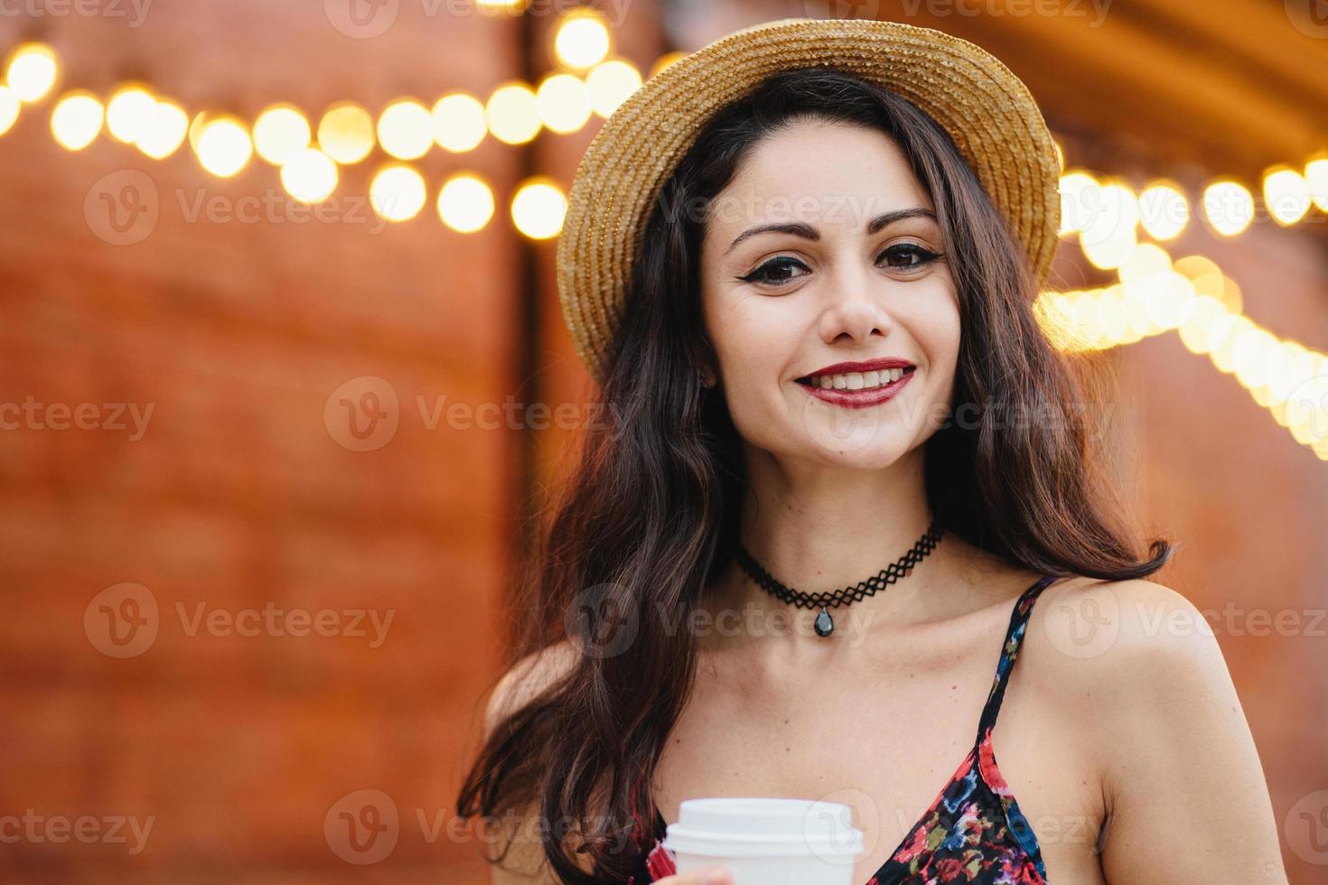 giovane modella con capelli lunghi scuri, occhi nocciola e labbra rosse, indossa collana, cappello estivo e vestito con un'espressione deliziosa mentre posa al caffè all'aperto. concetto di persone, riposo, emozioni foto