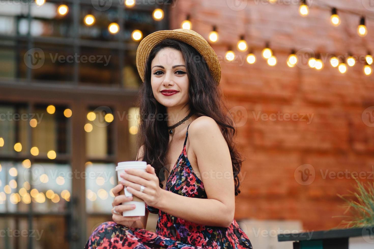 scatto all'aperto di una bella donna con i capelli lunghi scuri, con una pelle sana e pura, con indosso un cappello e un vestito di paglia, che beve tè caldo mentre è seduto sulla terrazza di un'accogliente caffetteria. bella donna che riposa al caffè foto