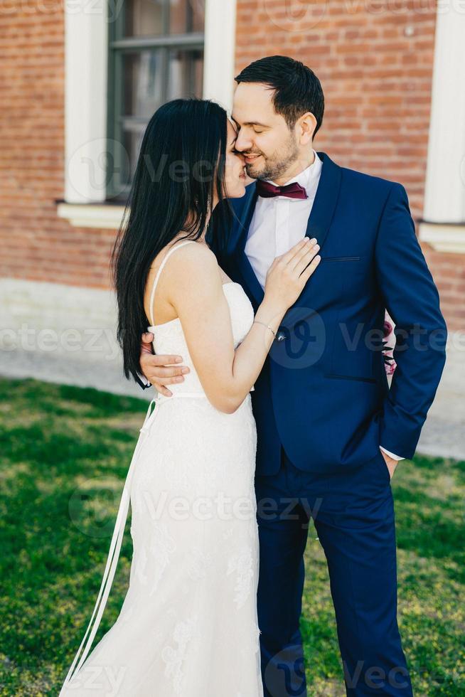 concetto di persone, relazioni e matrimonio. la sposa e lo sposo appena sposati si baciano appassionatamente, esprimono emozioni, amore e sentimenti reali. splendida coppia sposata posa all'aperto sull'erba verde. foto