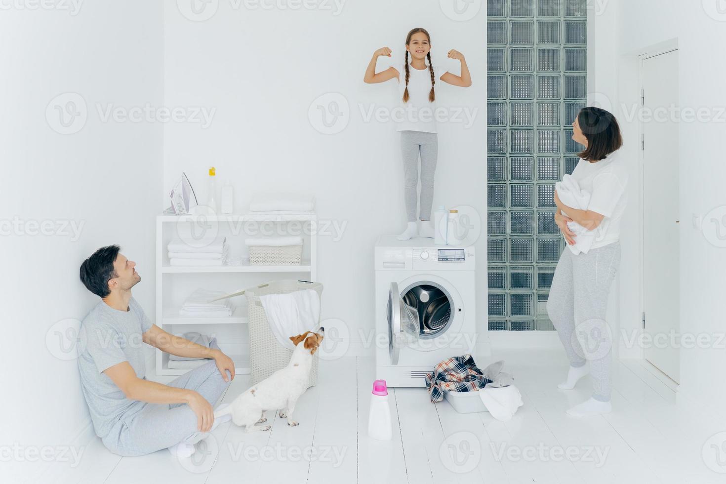 la foto di una scolaretta felice posa sopra la lavatrice, mostra i muscoli, alza le braccia, pronta ad aiutare i genitori con il bucato o il bucato. donna e uomo posano nella stanza di lavaggio con cane e bambino piccolo