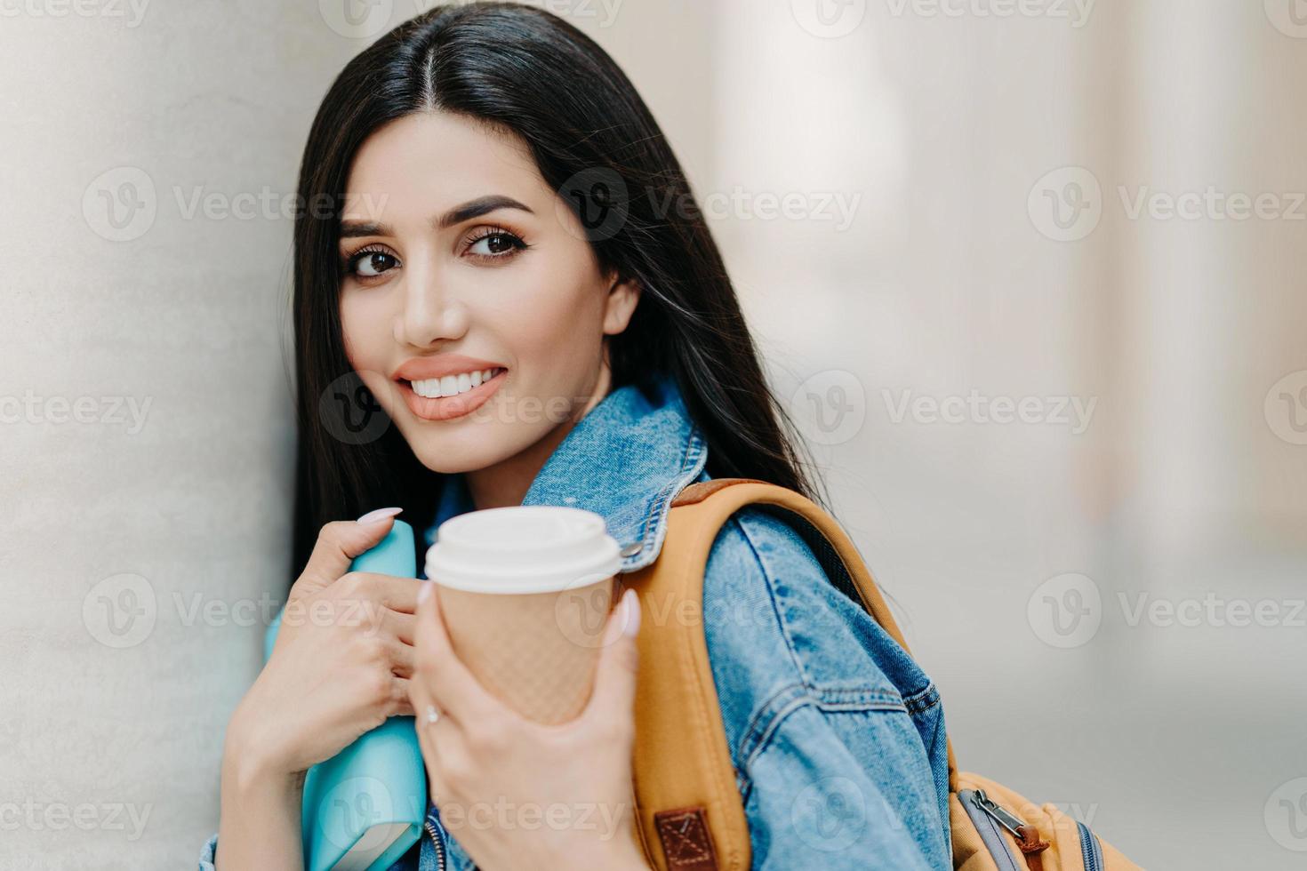 foto all'aperto di studentessa bruna con il trucco, capelli lunghi scuri, vestito con una giacca di jeans, tiene caffè da asporto, libro di testo, porta lo zaino, guarda da qualche parte in lontananza, passeggiate in città