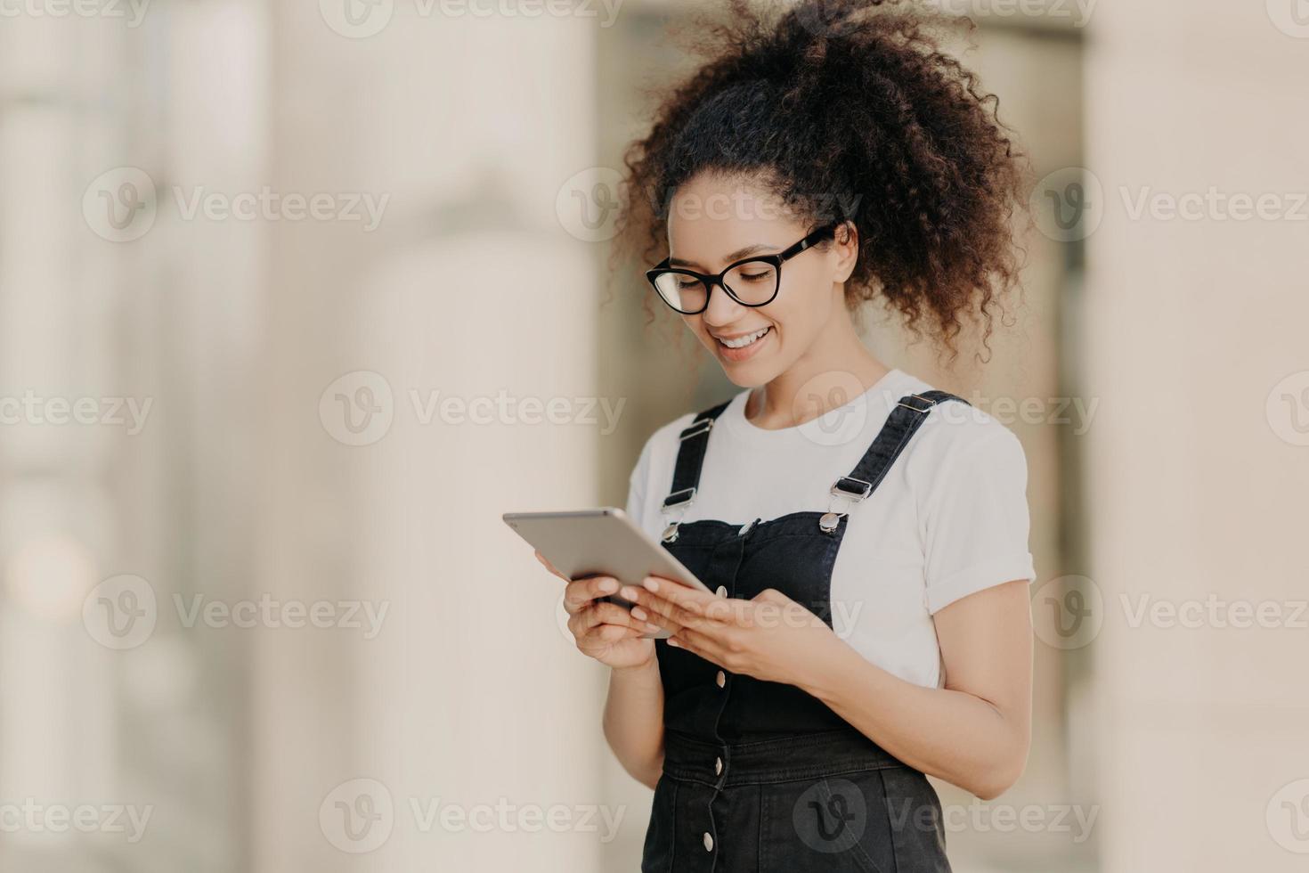 bella ragazza dai capelli ricci guarda lo schermo del tablet digitale con un sorriso, legge del testo o un libro elettronico, indossa occhiali per la correzione della vista, maglietta bianca e salopette, gode di internet ad alta velocità foto