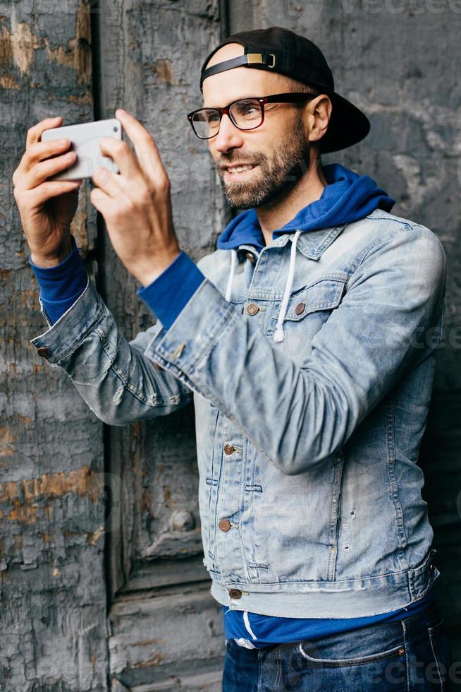 ritratto di ragazzo alla moda con la barba che indossa abiti alla moda che tiene il telefono cellulare facendo selfie essendo contento e sorridente con gioia nella fotocamera. maschio rilassato con aspetto attraente in posa a porte chiuse foto