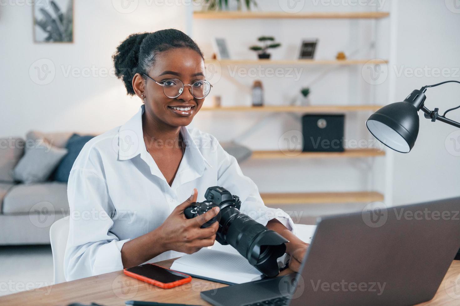 tenendo la fotocamera. la giovane donna afroamericana in camicia bianca è a casa foto