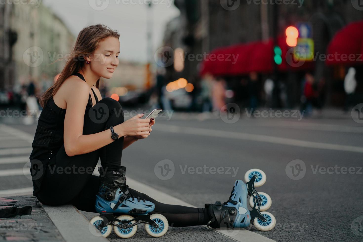 la foto all'aperto di una giovane donna sportiva e attiva vestita di abbigliamento attivo indossa i pattini a rotelle controlla il contenuto del messaggio sullo smartphone pone contro lo sfondo sfocato della città sull'asfalto conduce uno stile di vita sano