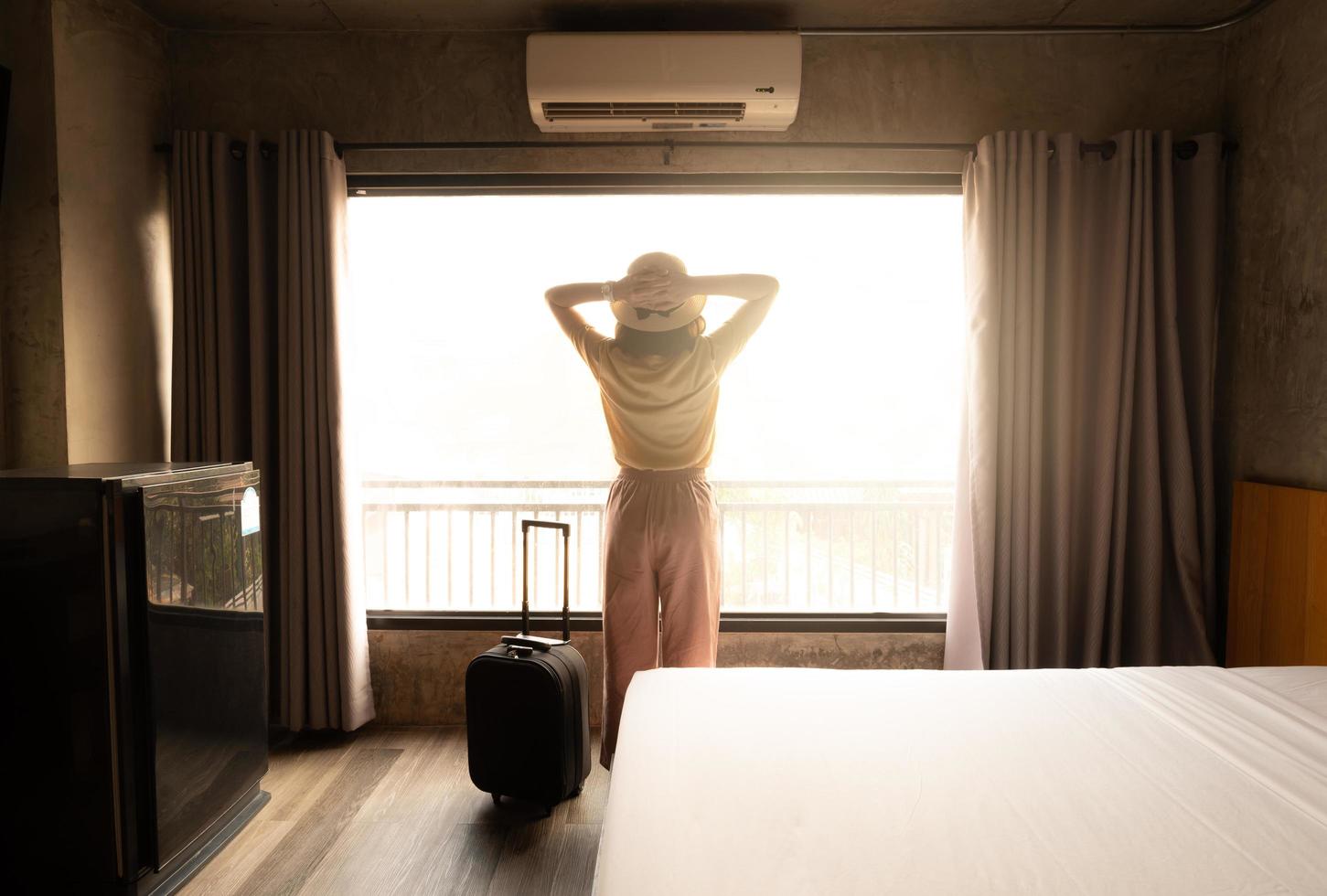 vista posteriore della donna turistica in piedi vicino alla finestra, guardando a una bella vista con i suoi bagagli nella camera da letto dell'hotel dopo il check-in. concettuale di viaggio e vacanza. foto
