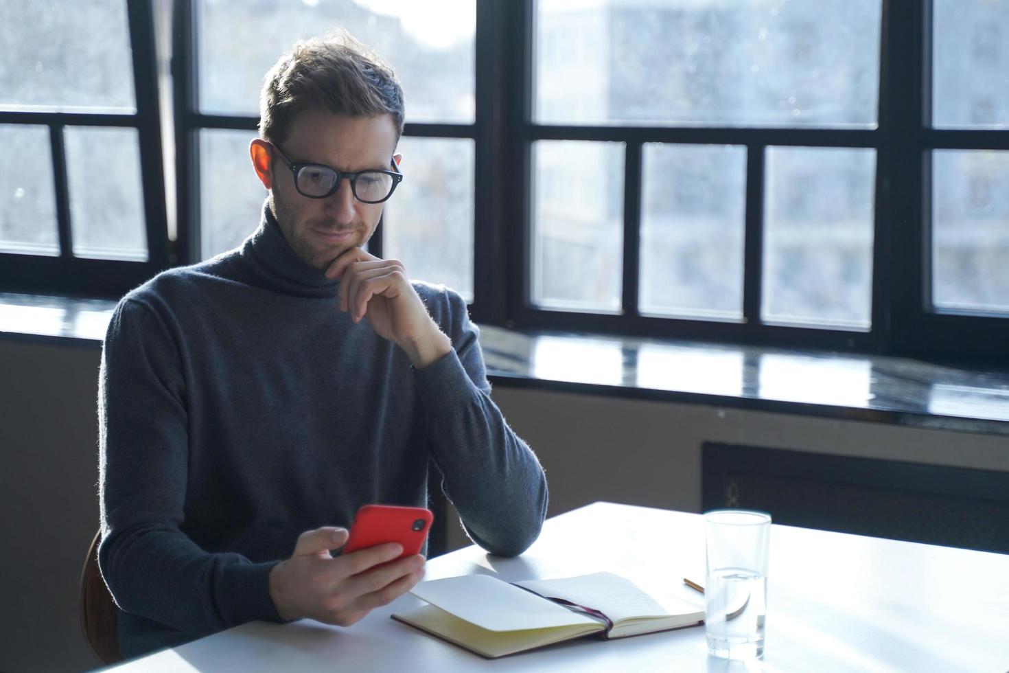 giovane uomo d'affari tedesco che legge notizie su Internet o controlla la posta elettronica sullo smartphone mentre è seduto alla scrivania foto