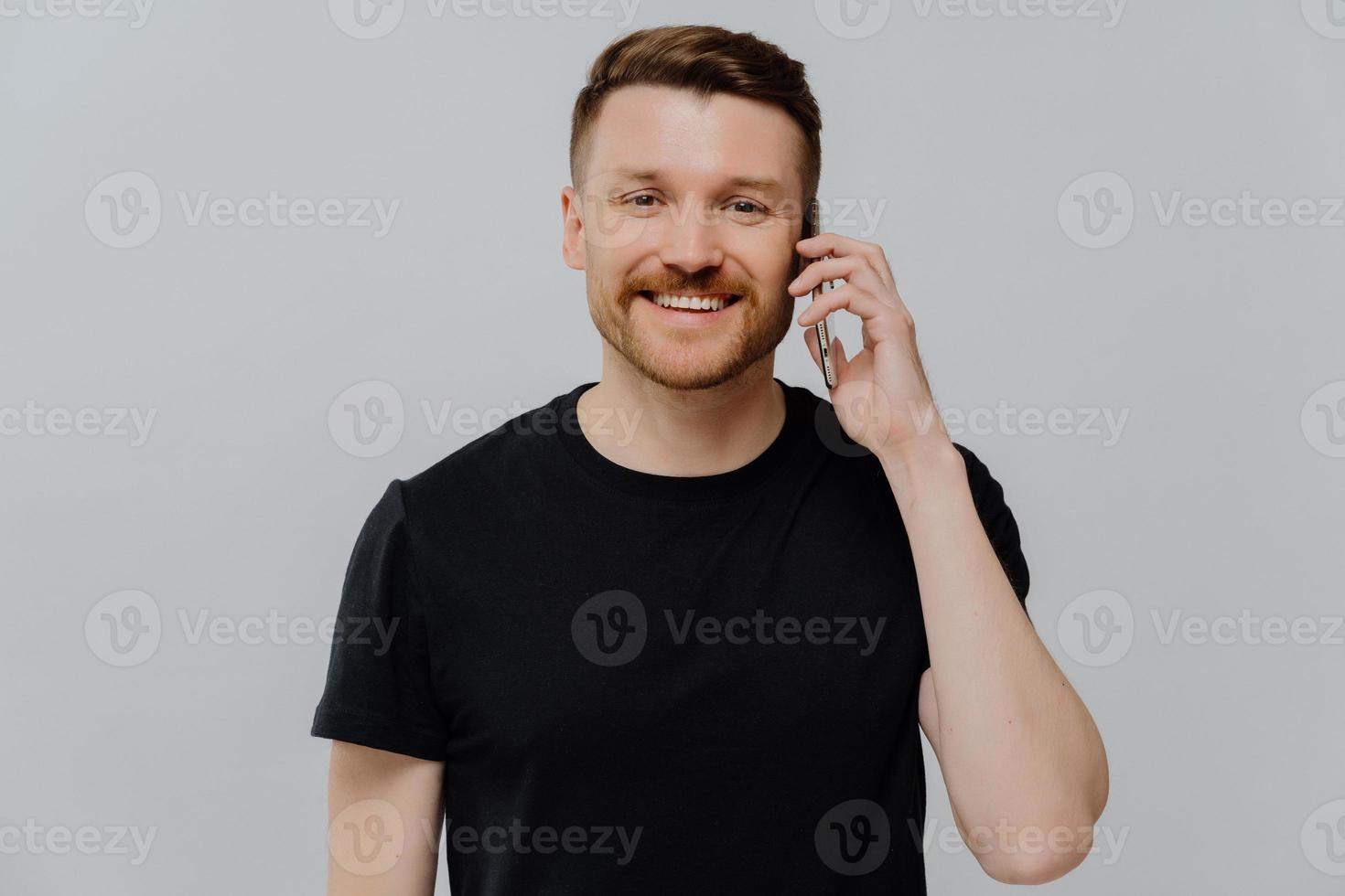 l'uomo adulto bello fa la chiamata tiene lo smartphone vicino all'orecchio gode di una piacevole conversazione con un amico vestito con una maglietta nera casual isolata su sfondo blu felice di sentire la voce delle amiche. foto