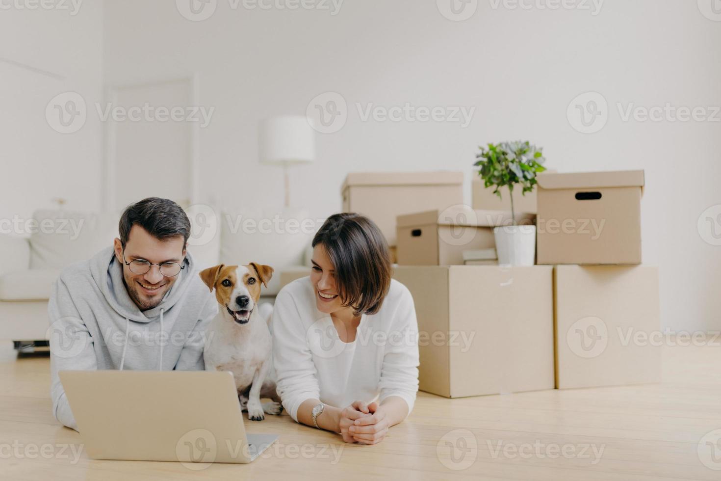 la coppia di famiglia si prende una pausa nel loro nuovo appartamento dopo essersi trasferita, si sdraia sul pavimento e naviga in Internet tramite un computer portatile, posa in una spaziosa stanza luminosa con divano, lampada e una pila di scatole di cartone, animale domestico foto