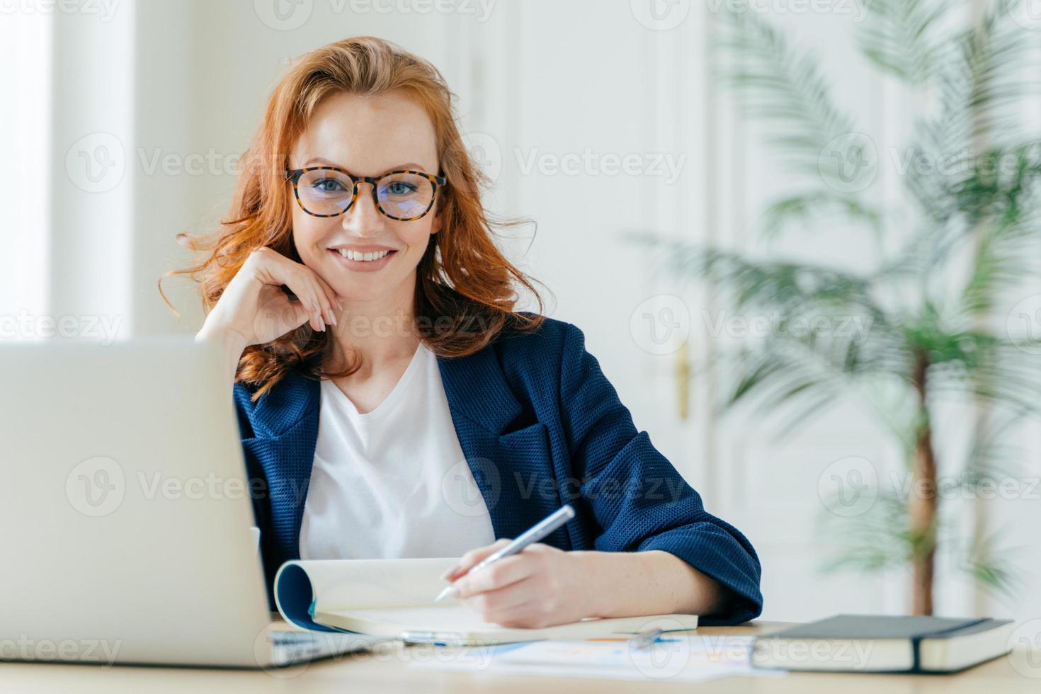 felice donna di successo scrive la lista di controllo nel blocco note, lavora su idee creative per il progetto di avvio, cerca le informazioni necessarie sul computer portatile, posa nello spazio di coworking, lavora come freelance foto