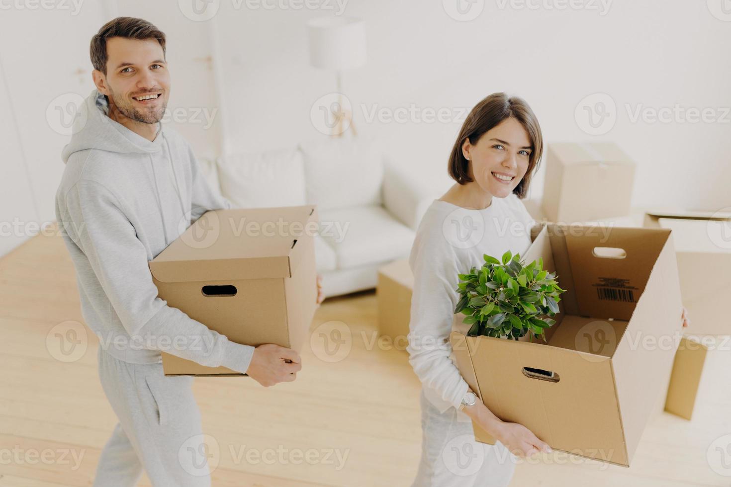 i proprietari di immobili di sesso femminile e maschile positivi posano con effetti personali in scatole di cartone, si trasferiscono nel proprio appartamento, sorridono piacevolmente, posano in un soggiorno bianco. trasloco, pianificazione, trasloco foto