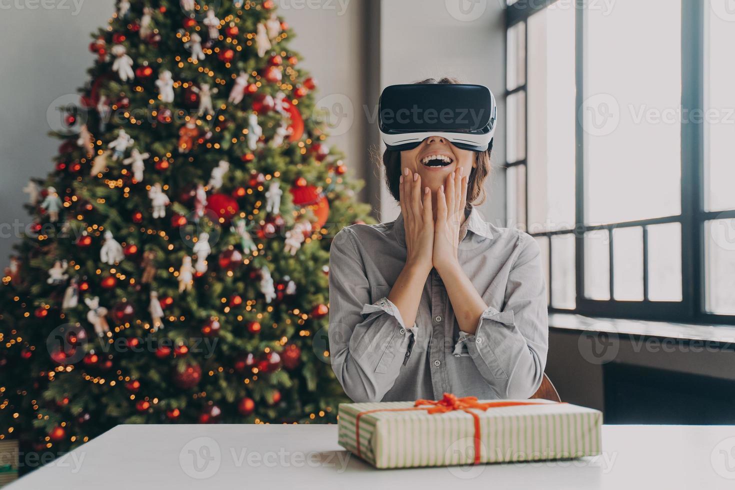 stupita giovane donna eccitata che indossa occhiali per realtà virtuale seduta contro l'albero di Natale foto