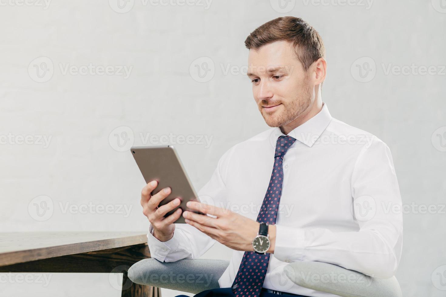 manager esecutivo maschile in camicia bianca e cravatta, tiene in mano un tablet, cerca le informazioni necessarie in internet, connesso al wifi in ufficio. concetto di persone, affari e tecnologie moderne foto