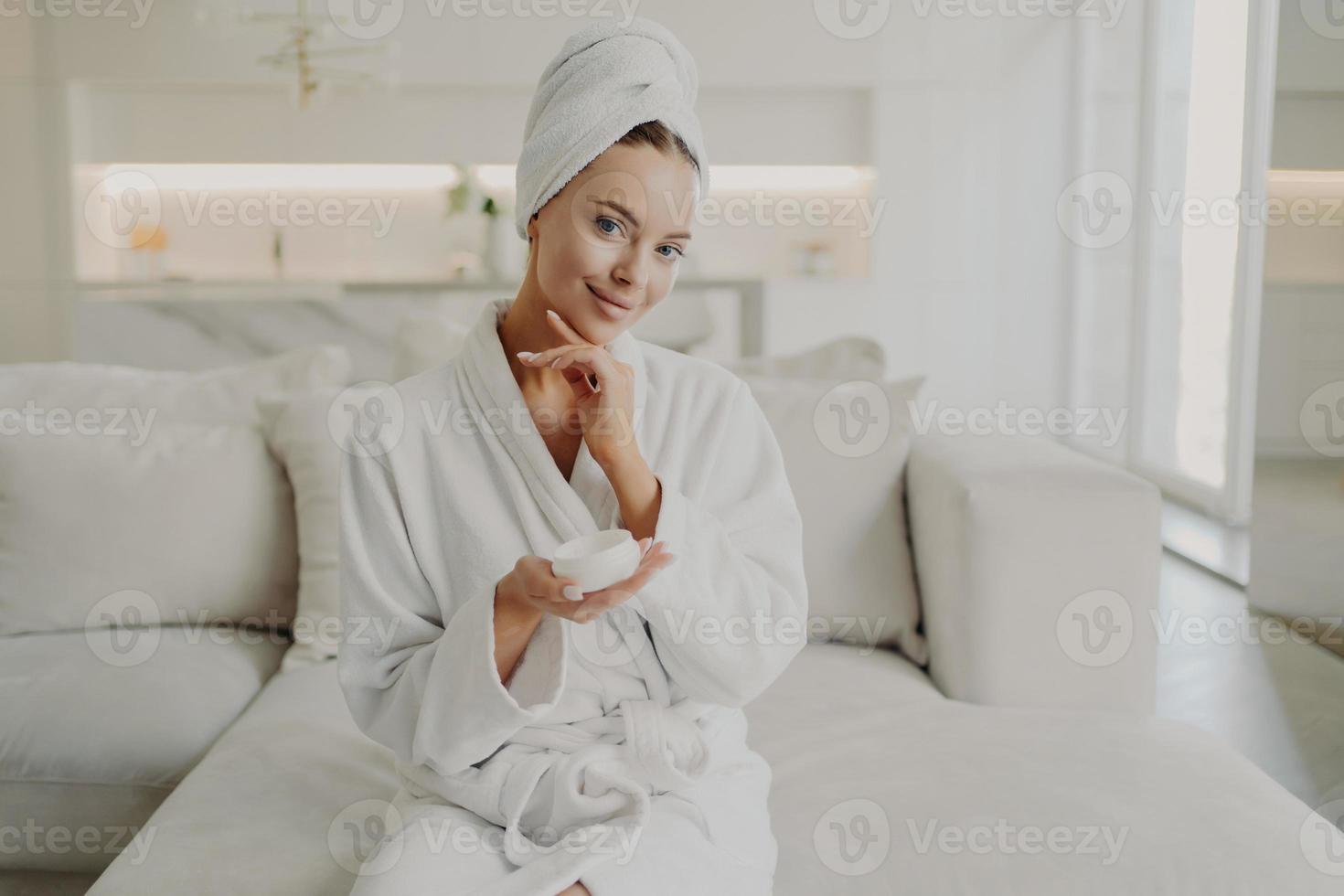 giovane bella donna che tiene un prodotto cosmetico e sorride mentre si rilassa sul divano dopo aver fatto la doccia a casa foto