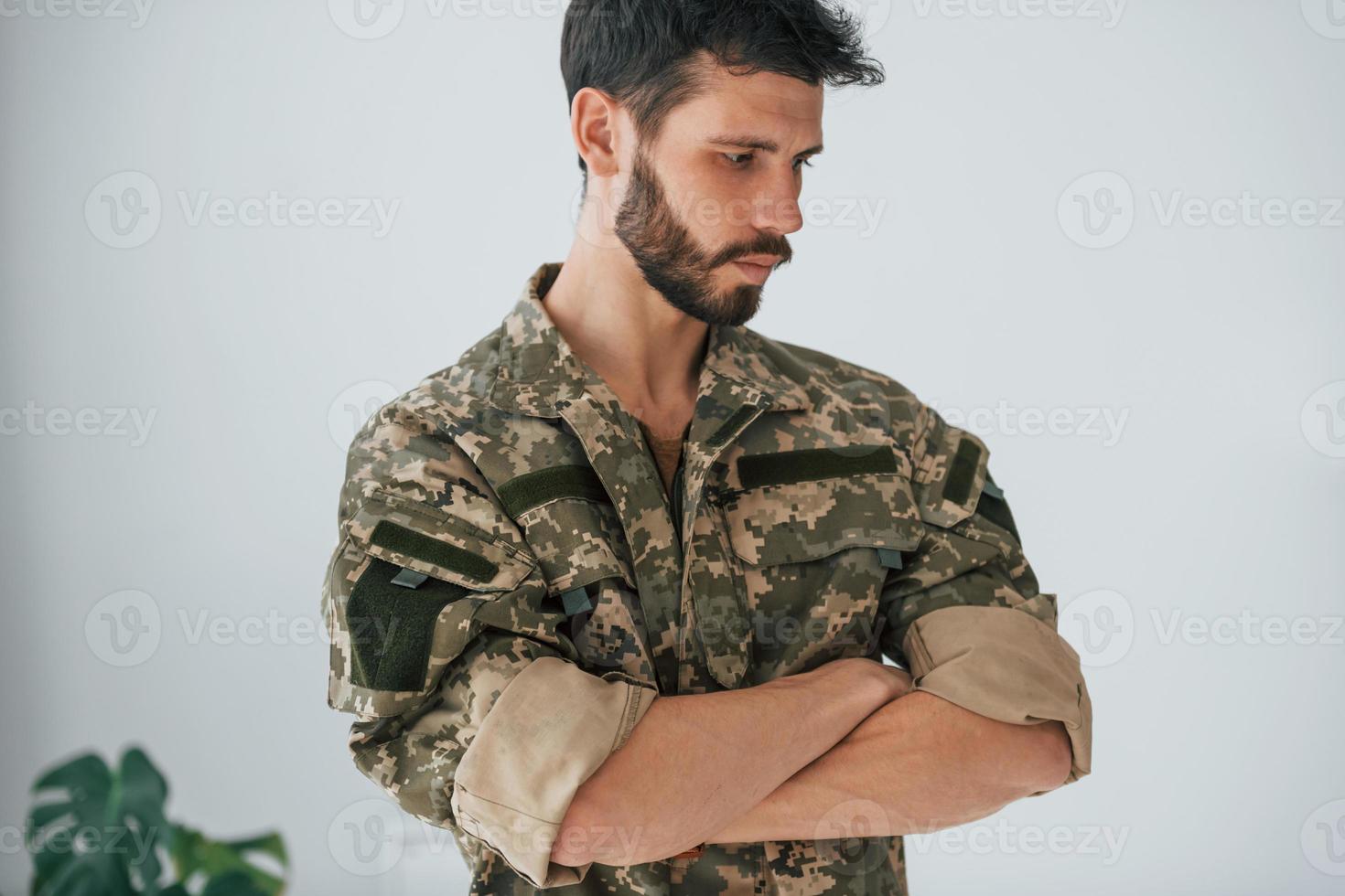 braccia incrociate. il soldato in uniforme è in piedi al chiuso contro il muro bianco foto