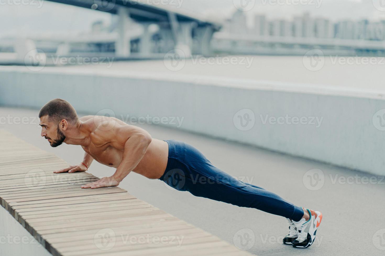il tiro orizzontale dell'atleta muscolare l'uomo fa esercizio di push up ha un corpo forte in posa in plank all'aperto indossa pantaloni sportivi e scarpe da ginnastica concentrate a distanza. concetto di forza e motivazione foto