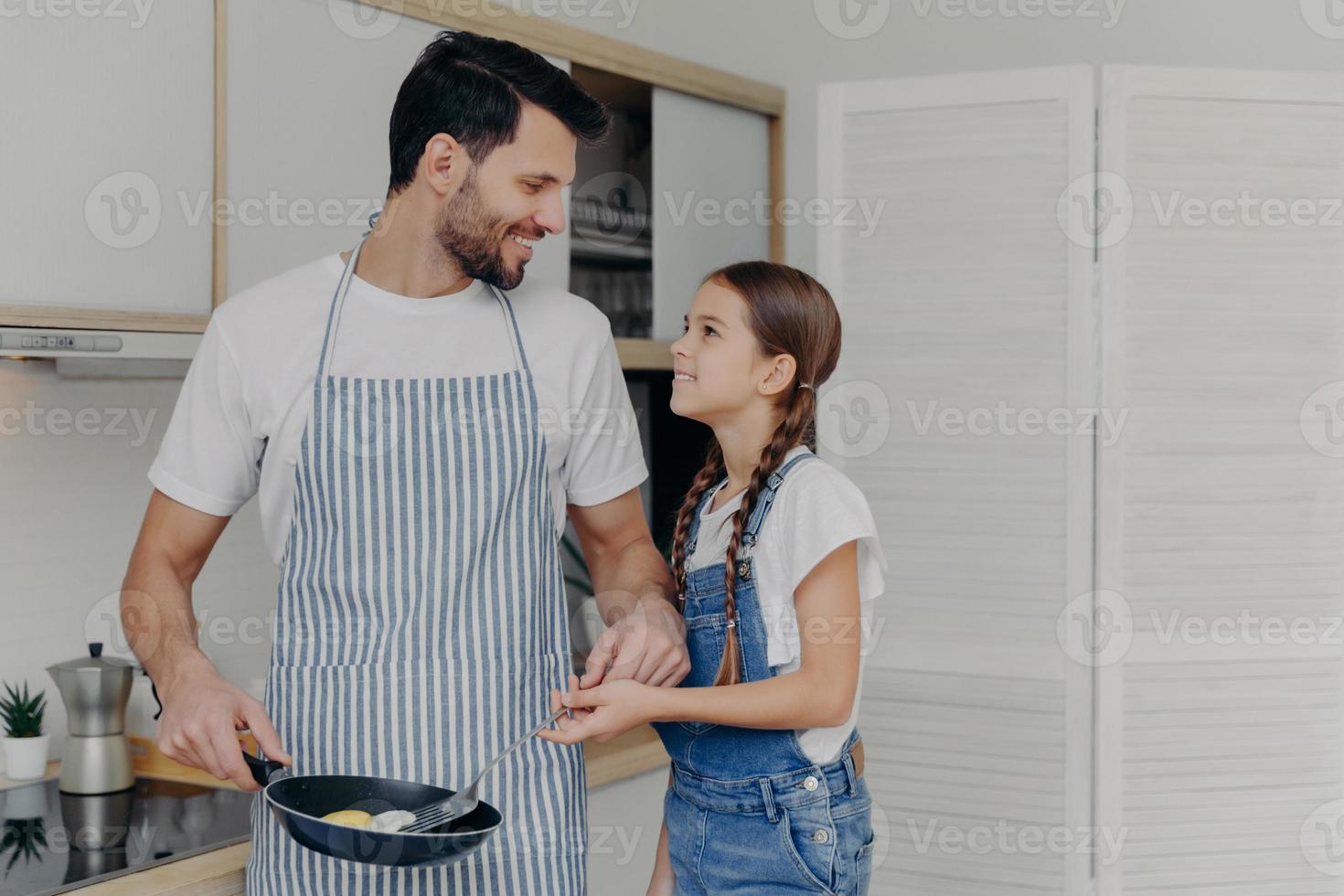 padre affettuoso in apton frigge le uova, insegna alla figlia piccola come cucinare, posa insieme in cucina, prepara una deliziosa colazione, guardati con un sorriso. figli, paternità, vita domestica foto