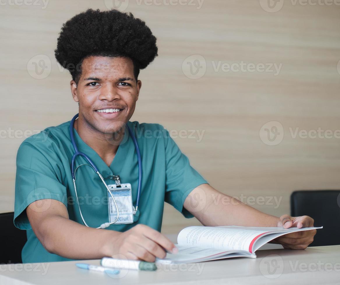 Ritratto di studente di medicina seduto alla scrivania guardando la fotocamera sorridente. il giovane medico afroamericano legge il libro e i documenti che si preparano per gli esami. professione sanitaria e professione medica. foto