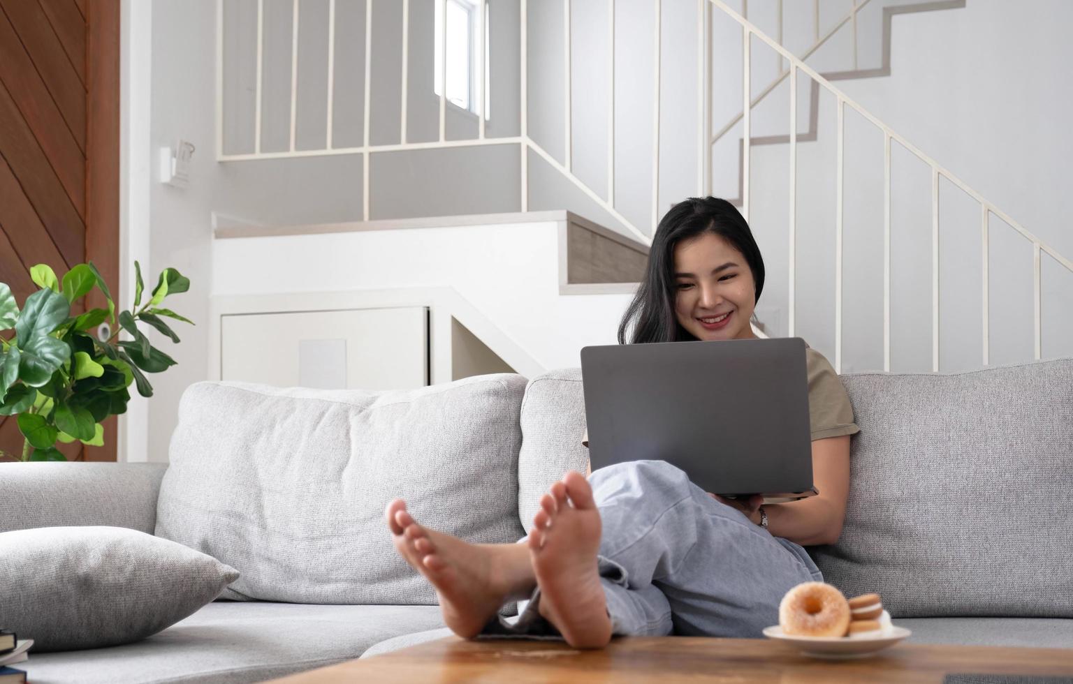 giovane donna asiatica freelance che lavora al computer portatile controllando i social media mentre è sdraiato sul divano quando si rilassa nel soggiorno di casa. foto