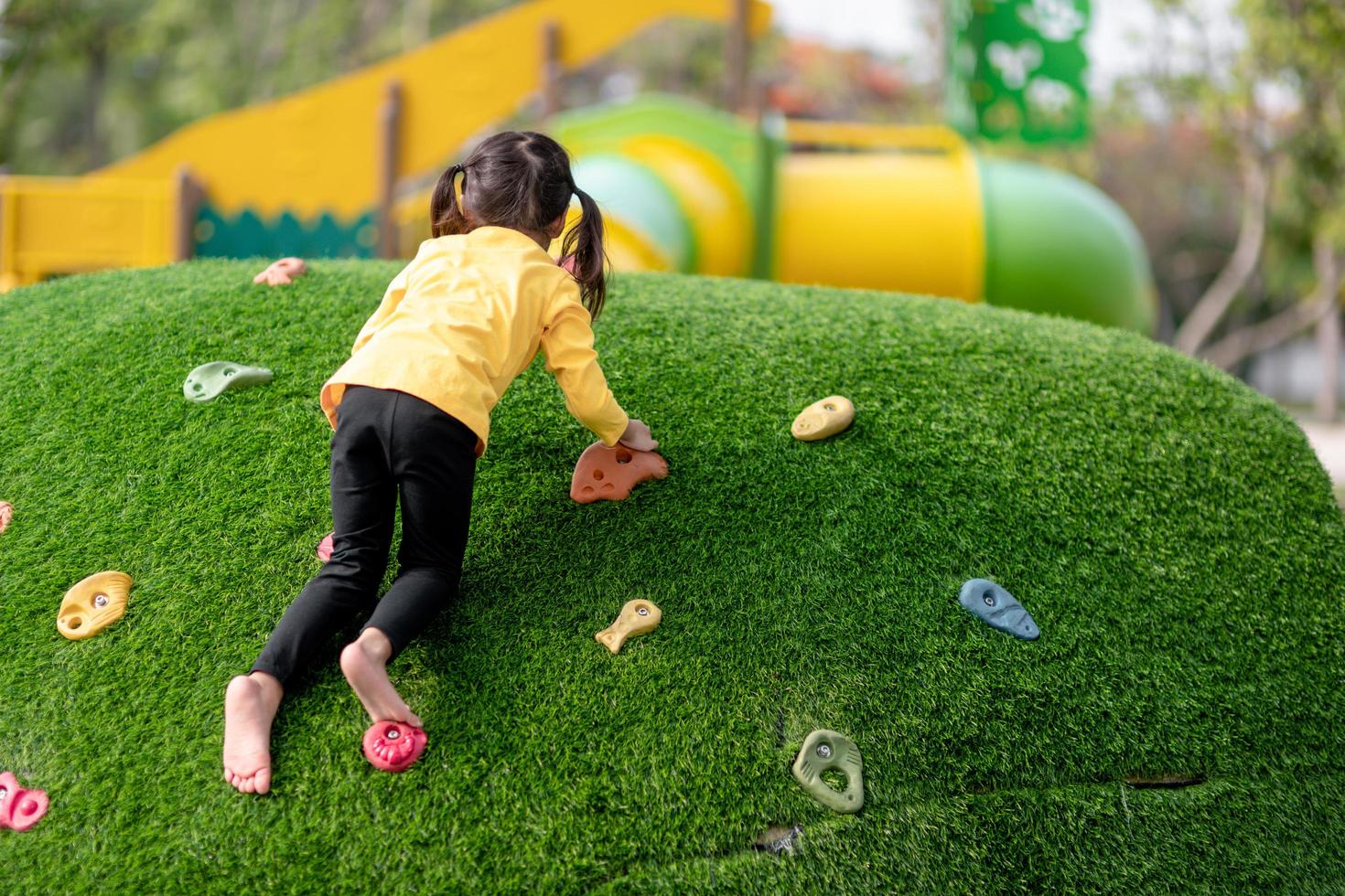 carina ragazza asiatica che si diverte cercando di arrampicarsi su massi artificiali nel cortile della scuola, bambina che si arrampica sulla parete rocciosa, coordinazione occhio-mano, sviluppo delle abilità foto