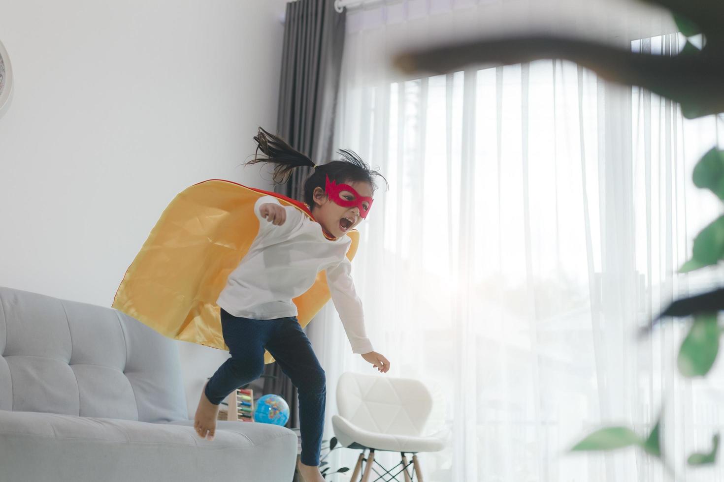 bambina che salta sul divano del soggiorno di casa indossando un vestito da supereroe improvvisato, giocando divertendosi con giochi energici, al chiuso. scoperta attività per bambini vacanza stile di vita. foto