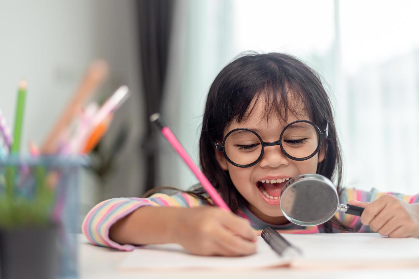 studentessa asiatica che scrive con una lente d'ingrandimento guardando la telecamera. dolci bambini seduti nel soggiorno di casa. concetto di attività familiare. foto