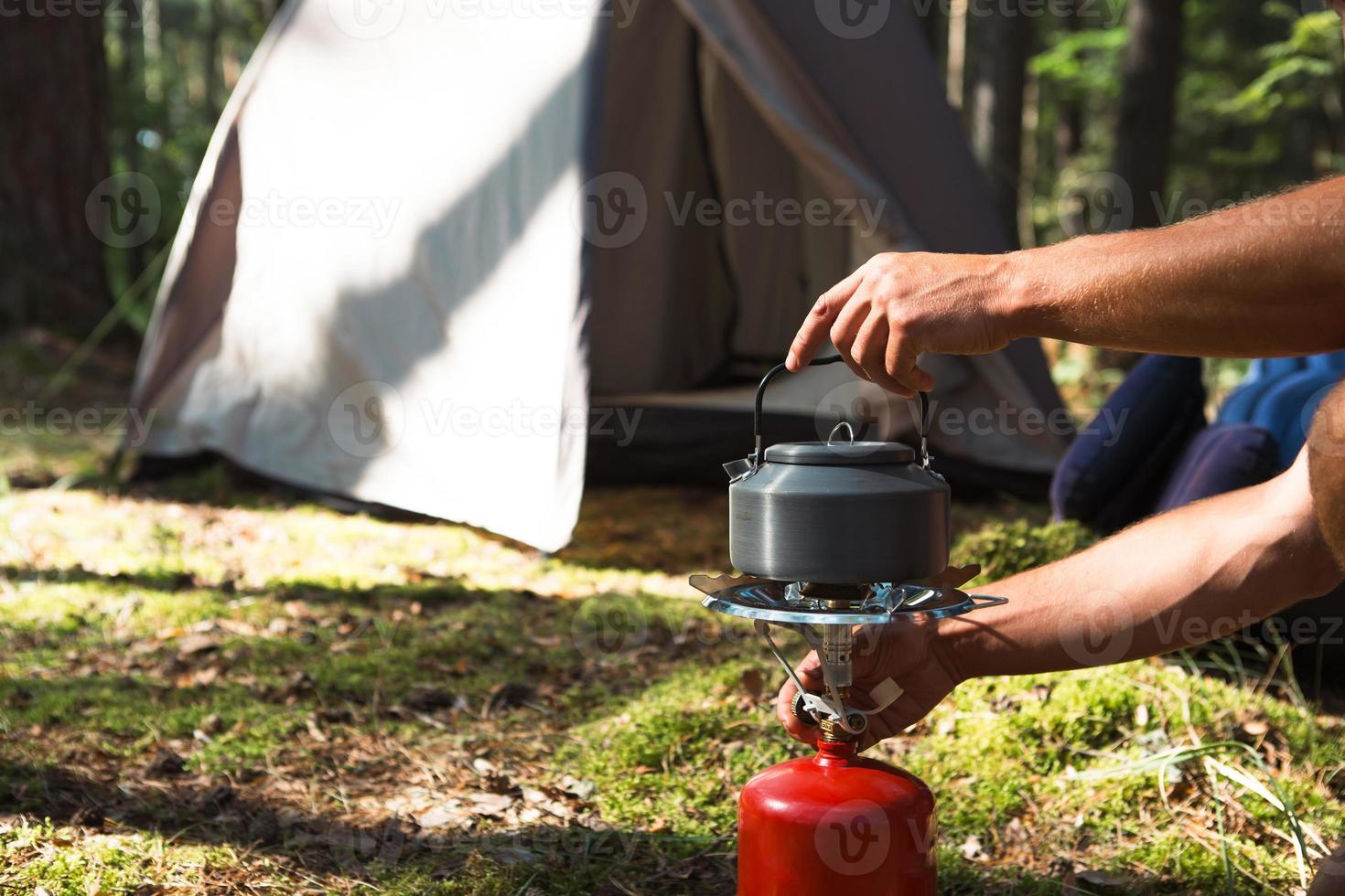 cucinare, riscaldare un bollitore turistico su un bruciatore a gas portatile con una bombola di gas rossa. campeggio, un uomo prepara la colazione all'aperto. attività estive all'aperto foto