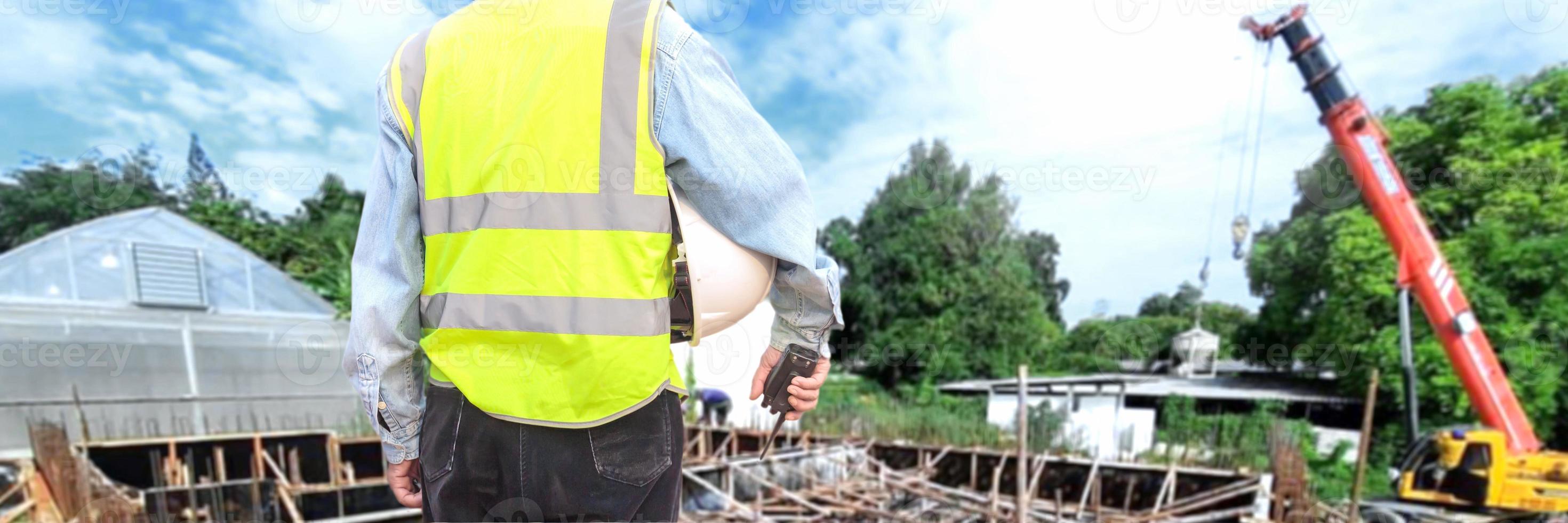 un ingegnere o un lavoratore che indossa un giubbotto di salvataggio tiene in mano un walkie-talkie e un berretto bianco. per la sicurezza lavorando sullo sfondo del cantiere foto