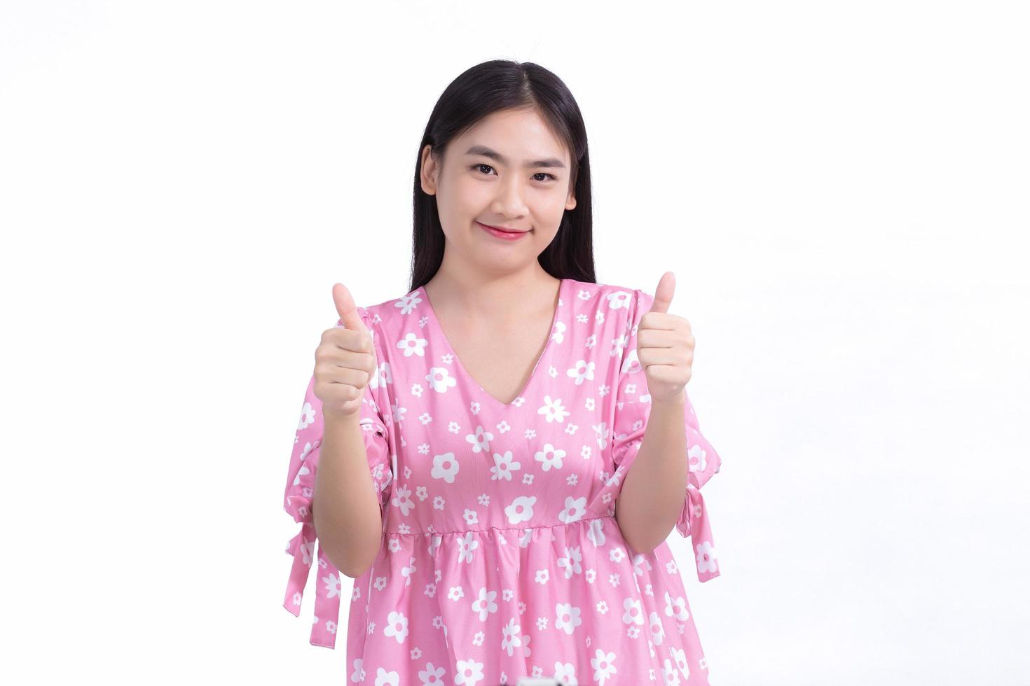 bella donna asiatica con capelli lunghi neri in un vestito rosa, sorridente di buon umore, pollice in alto e presentando qualcosa su uno sfondo bianco. foto