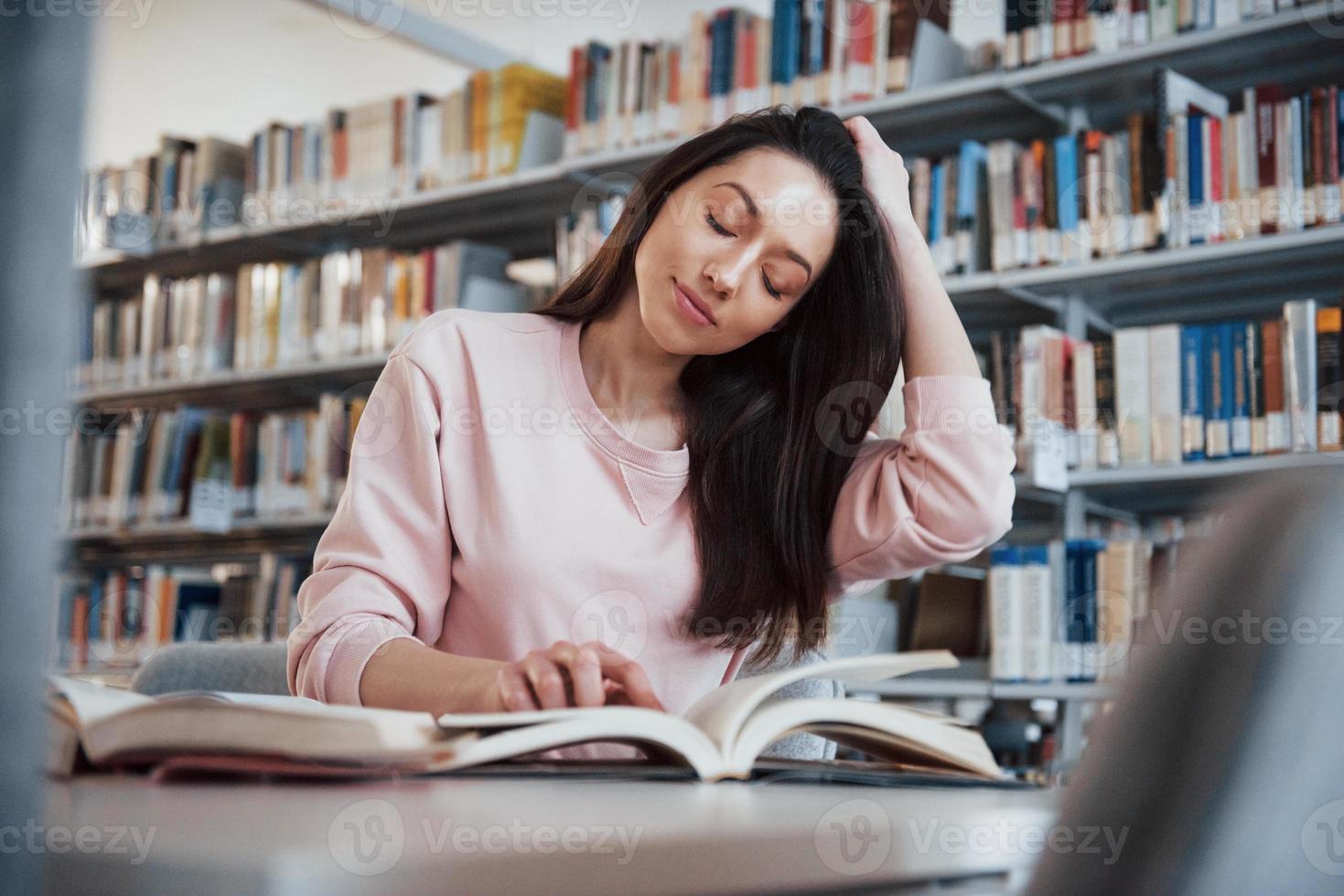 toccando i capelli. ragazza bruna in abiti casual divertendosi nella biblioteca piena di libri foto