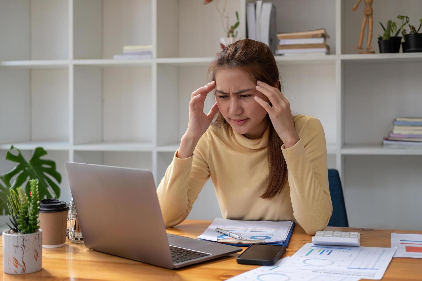 la donna che lavora ha mal di testa, la giovane donna d'affari asiatica che lavora su un laptop e le scartoffie è stressata ha mal di testa e pensa intensamente in ufficio foto