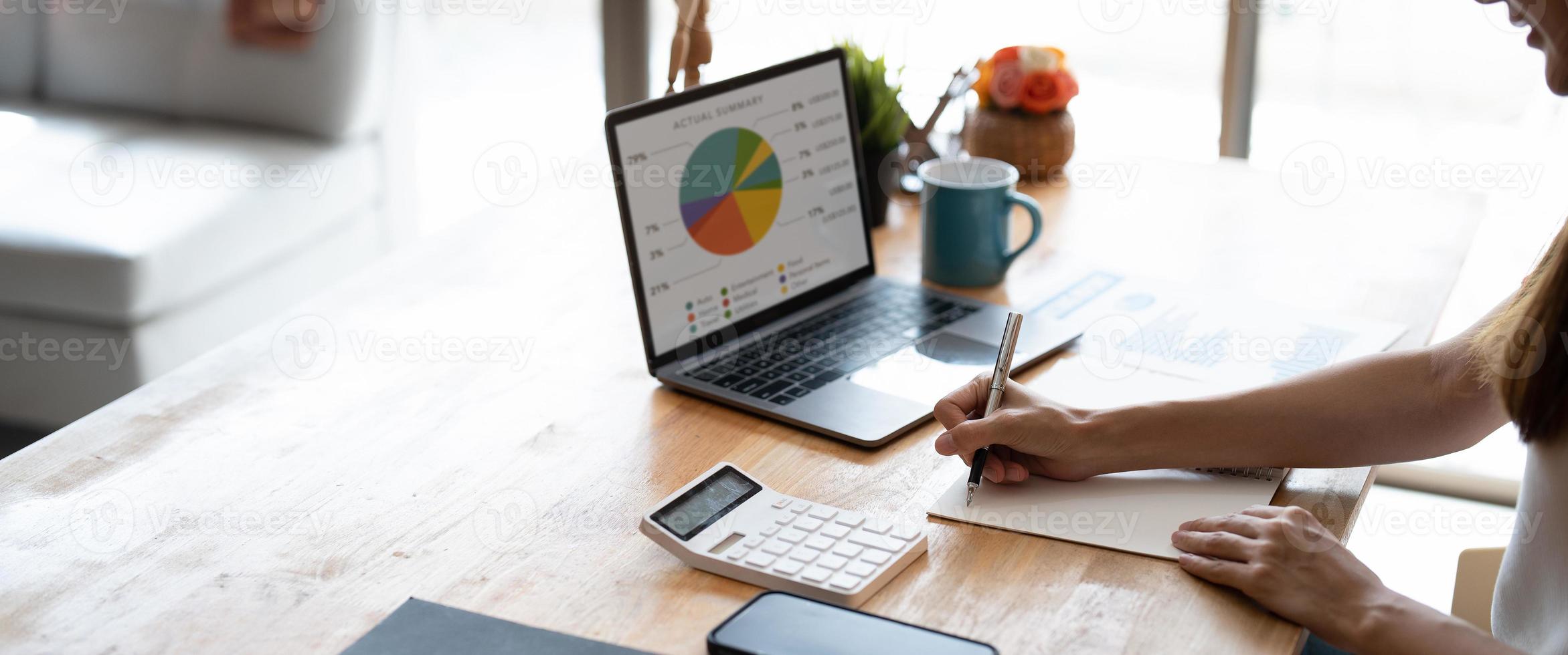 primo piano di mani femminili con computer portatile e penna per prendere appunti durante il calcolo con calcolatrice a casa foto