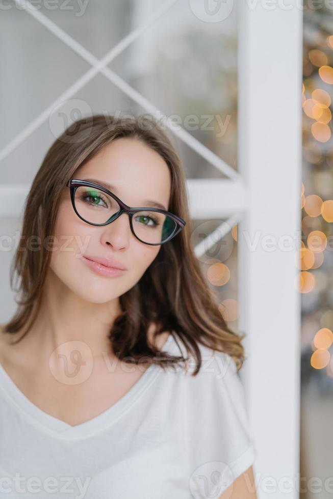 inquadratura verticale di una donna affascinante con occhiali da vista, vestita in modo casual, posa al chiuso contro l'albero di Natale, ha una pelle morbida e sana, gode del tempo libero nell'atmosfera domestica. persone e tempo libero foto