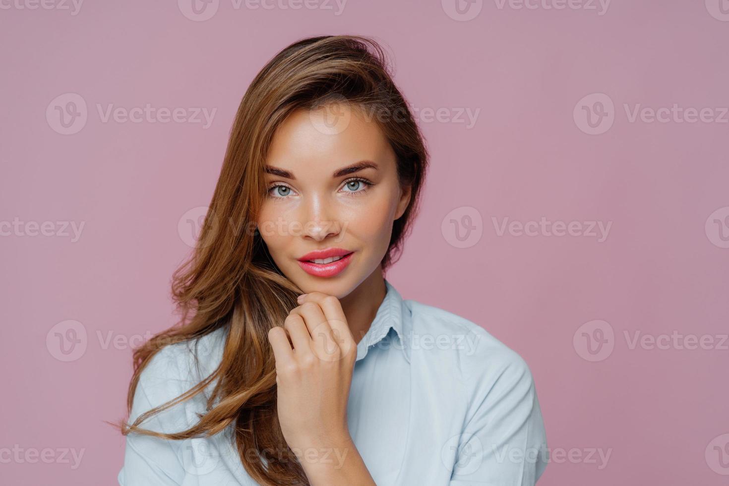 il ritratto di una bella donna dai capelli lunghi tocca il mento, ha gli occhi azzurri, guarda con sicurezza la fotocamera, indossa una camicia casual, ha il trucco per sembrare bella, posa su sfondo viola. donne, bellezza foto