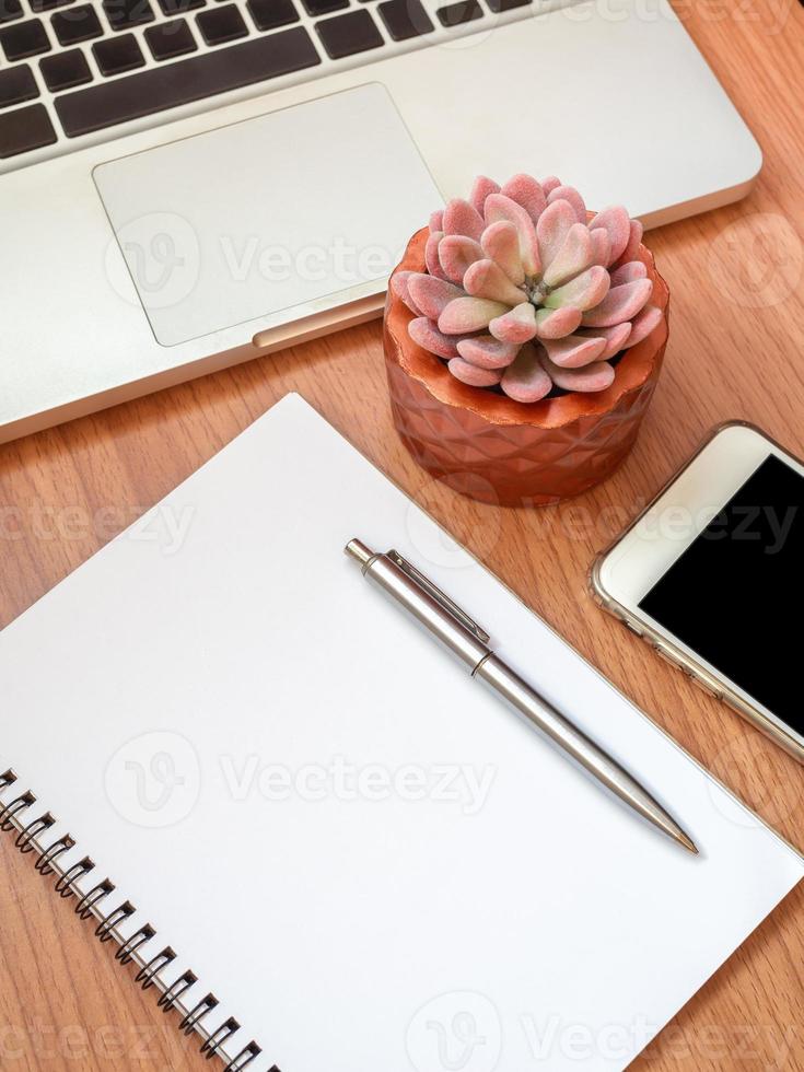 taccuino in bianco con smartphone, computer portatile e pianta succulenta in vaso di rame sul fondo della tavola di legno foto