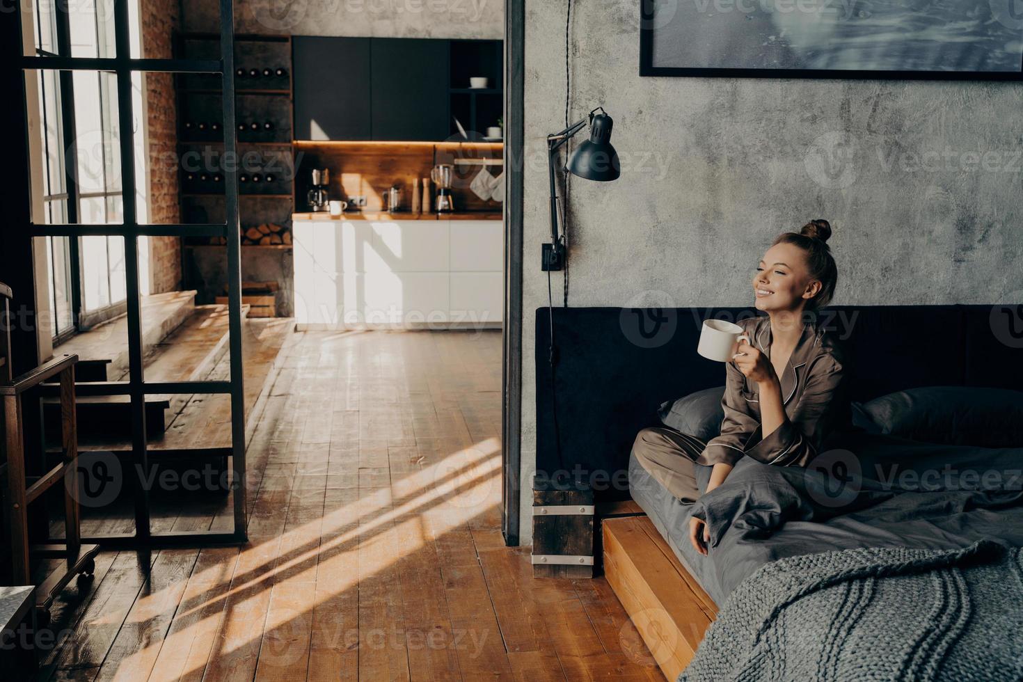 giovane ragazza attraente felice seduta sul letto nella posizione del loto e bere il caffè del mattino foto